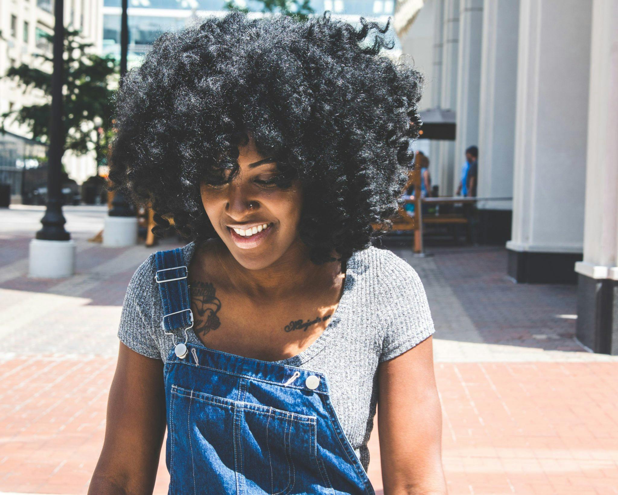 A young woman with curly hair smiles outdoors in an urban environment during the day.